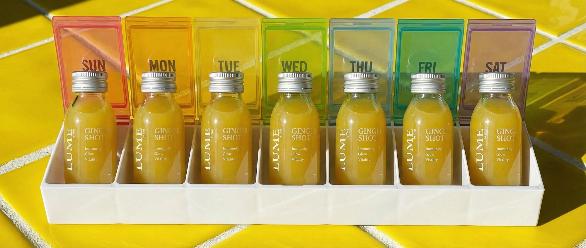 Six LUME ginger shot bottles in a daily pill tray labelled Sun to Sat — building a consistent daily wellness habit